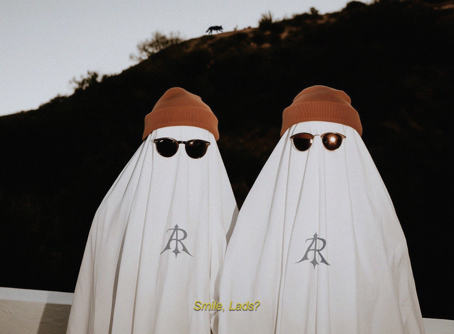 Two people wearing white ghost costumes with logos and sunglasses, standing against a dark background.